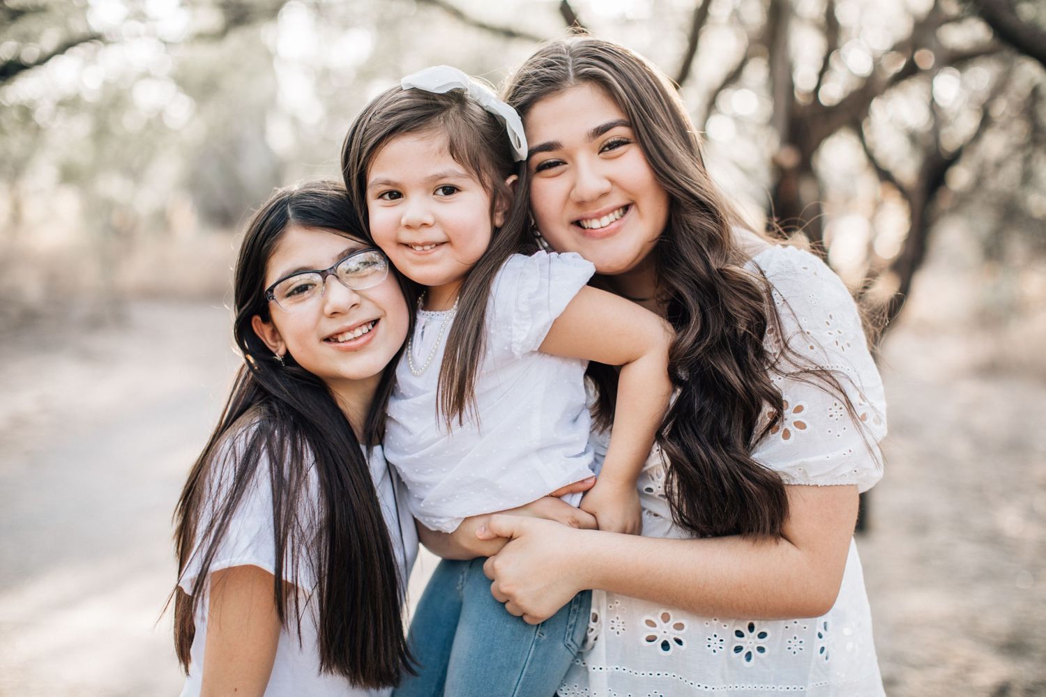 Tucson sibling portrait of three sisters smiling and hugging in soft natural light on a tree-lined path.