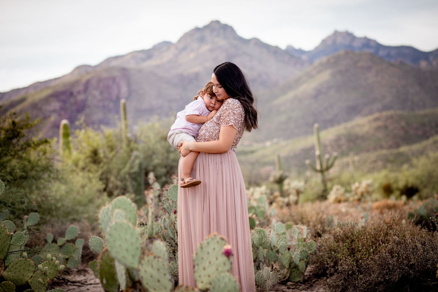 Tucson maternity session showing an expecting mother in a flowing pink dress surrounded by desert cactus and mountain views.