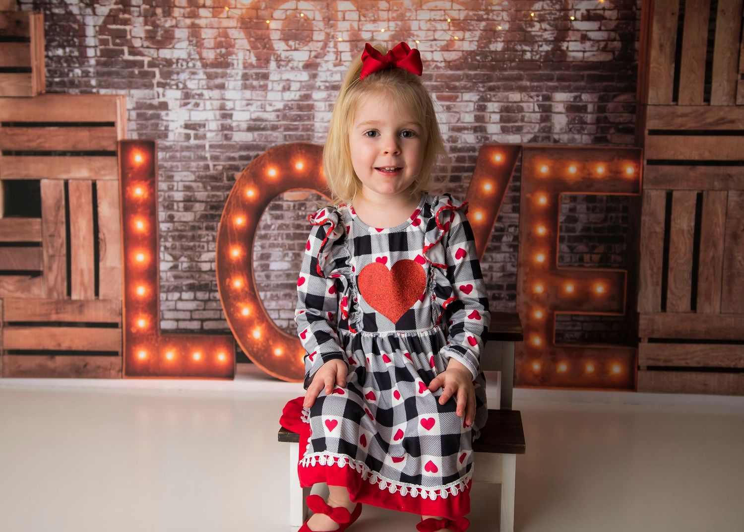 A child in a heart-print dress with pom-poms stands before illuminated letters and wooden crates.