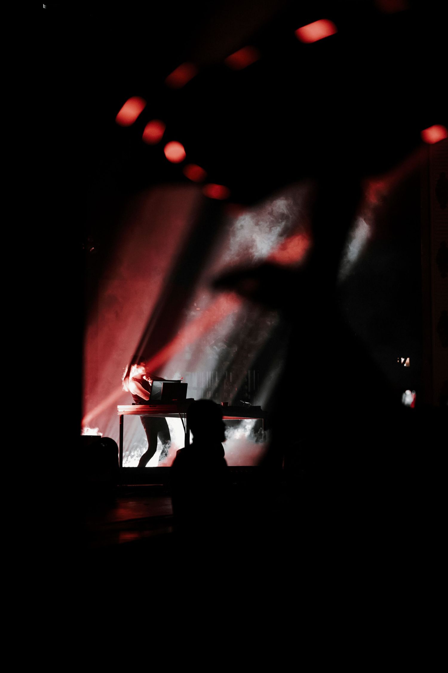 Atmospheric concert lighting creates moody silhouette of DJ equipment in red and white hues.