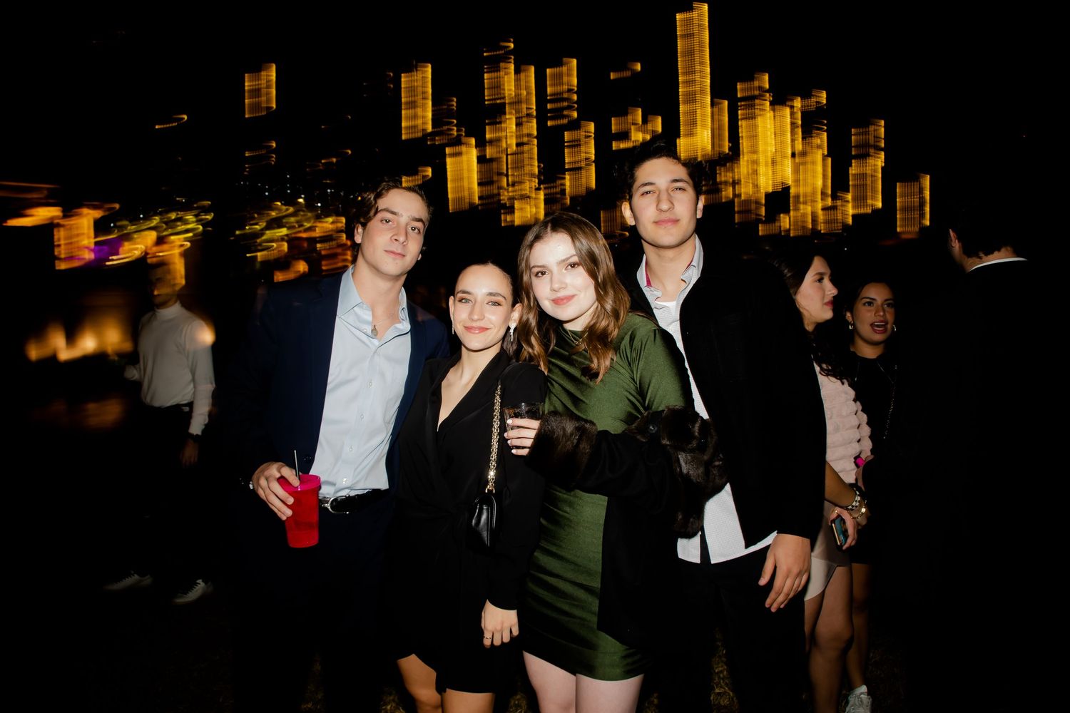 Four friends pose together at a rooftop night party with city lights glowing in the background.