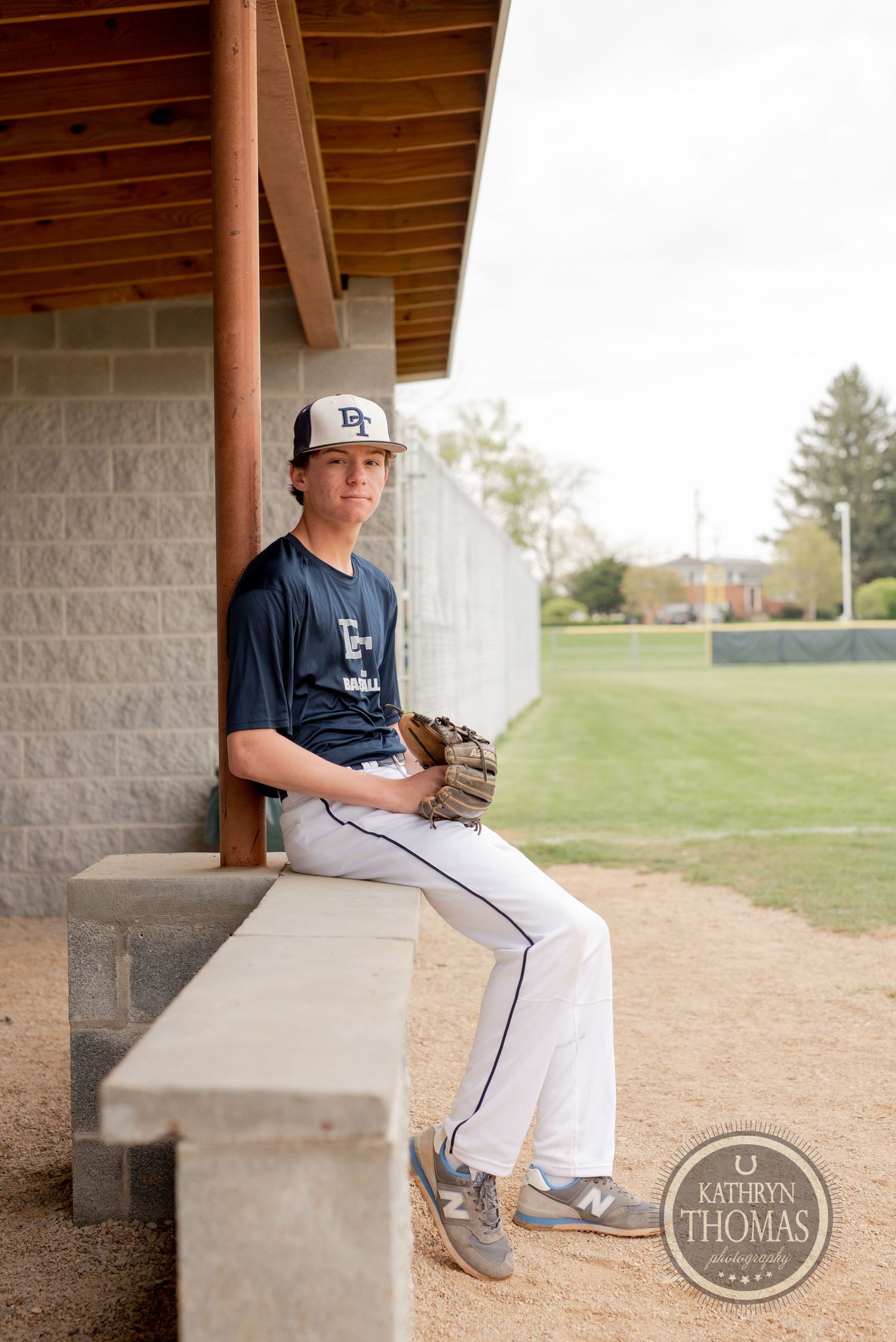 Baseball player sits in dugout wearing navy jersey and white pants with baseball glove.