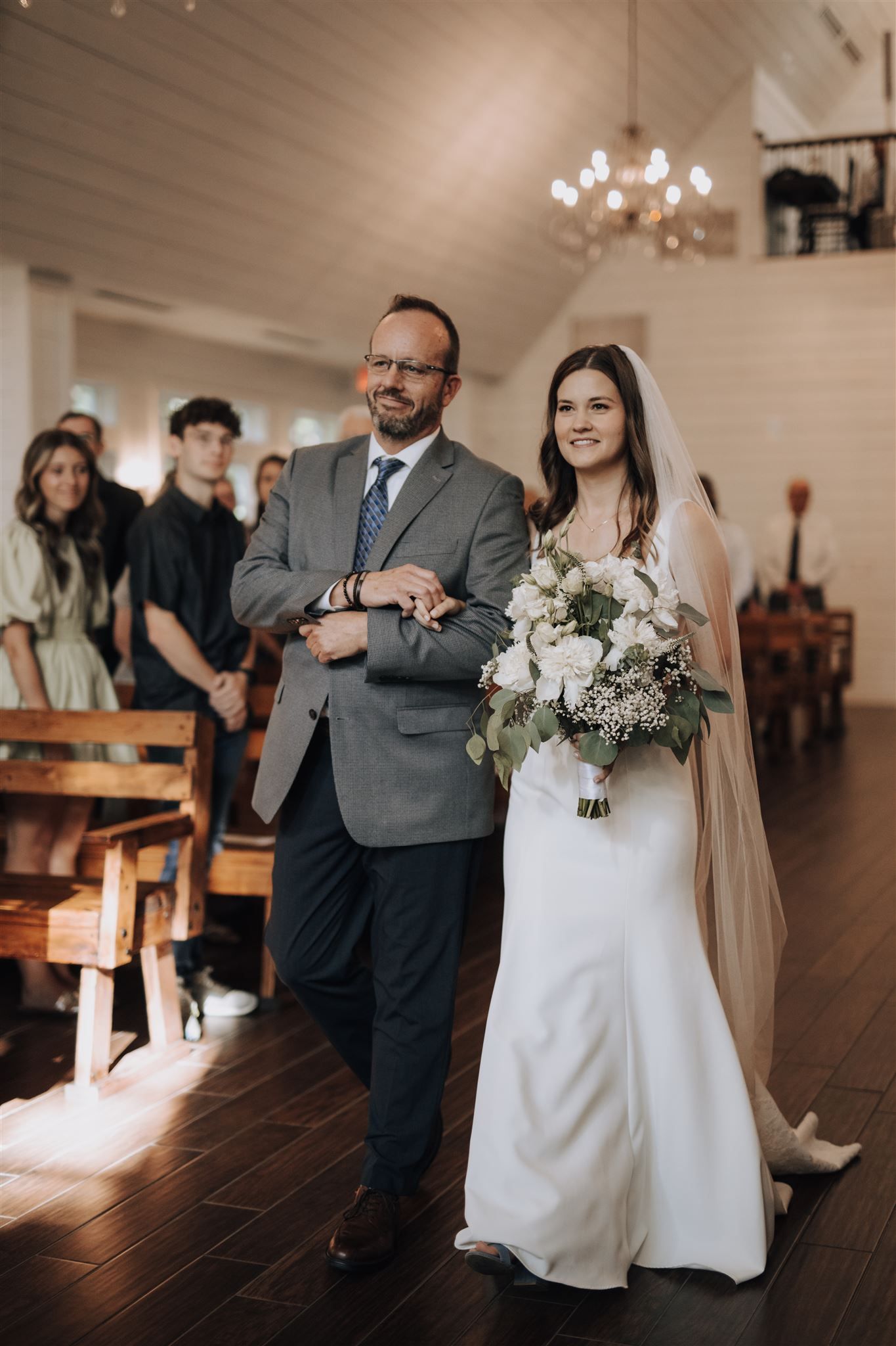 A bride in white gown walks down the aisle with her escort in a gray suit.