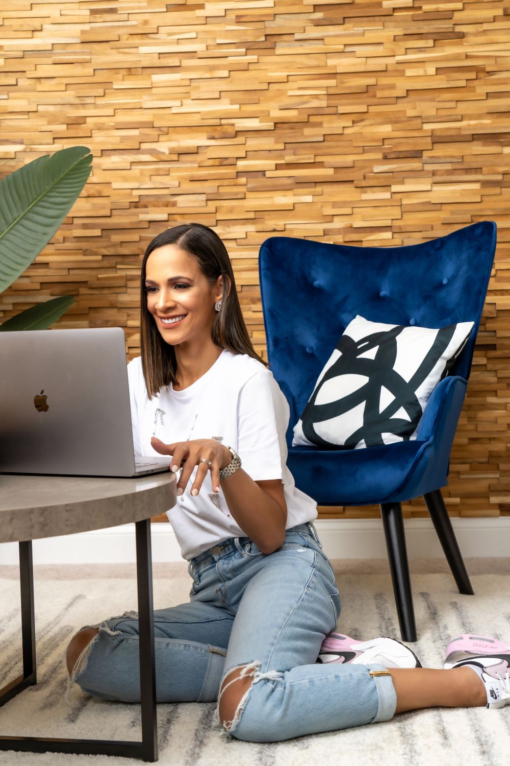 Woman in casual attire working on MacBook laptop while sitting on floor next to blue accent chair.