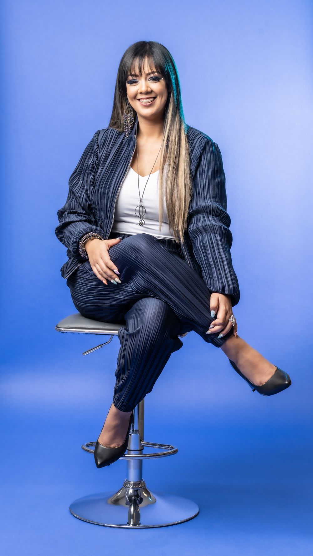 Portrait of person in pinstripe outfit sitting on chrome bar stool against blue backdrop.