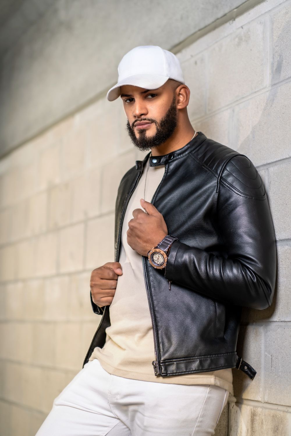 Fashion photo of person in black leather jacket and white baseball cap against concrete wall.