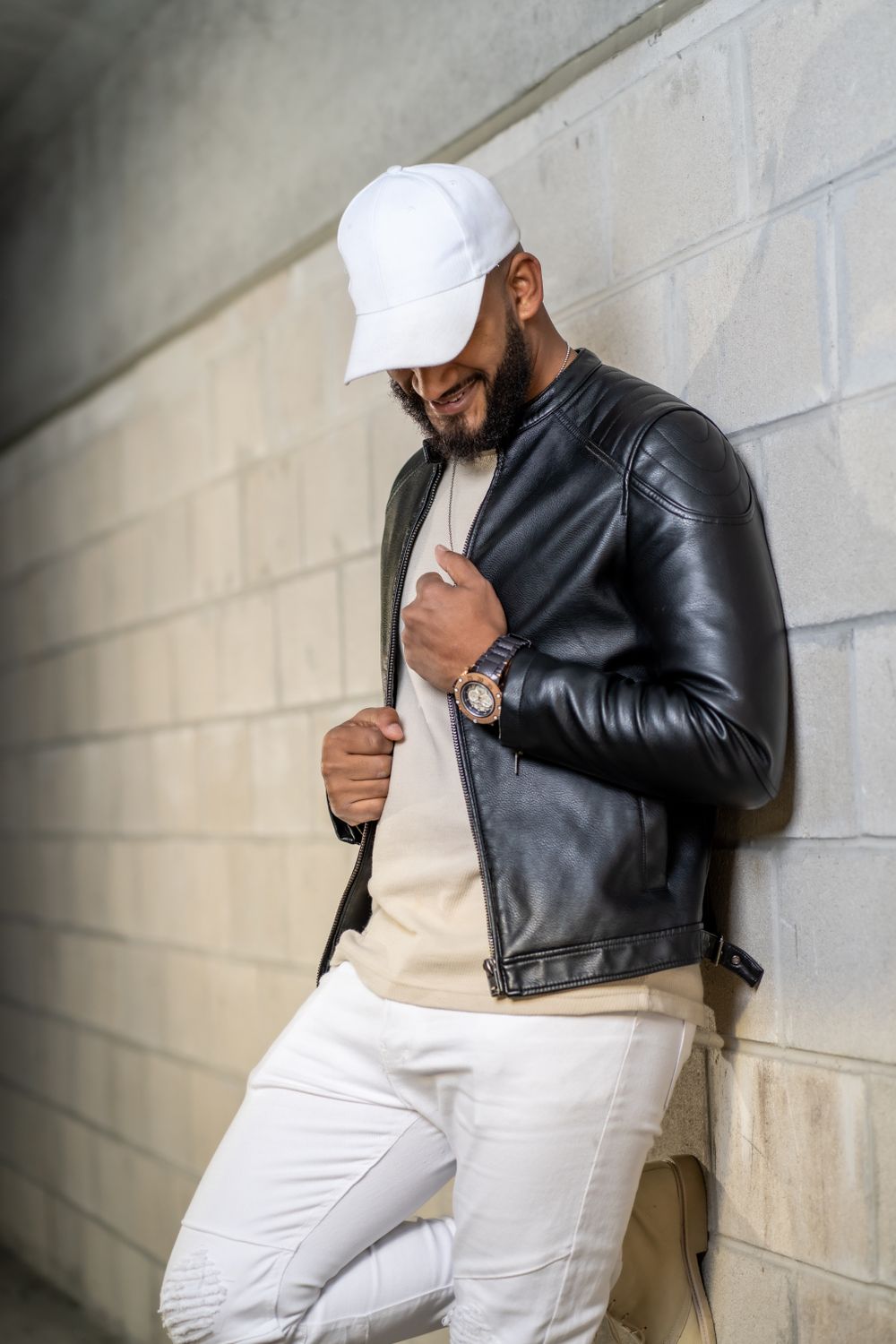 Person in leather jacket and white cap posing against concrete wall.