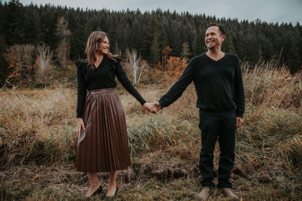 A smiling couple holds hands in a grassy autumn field, the woman in a pleated skirt and the man in dark clothing, with evergreens behind.