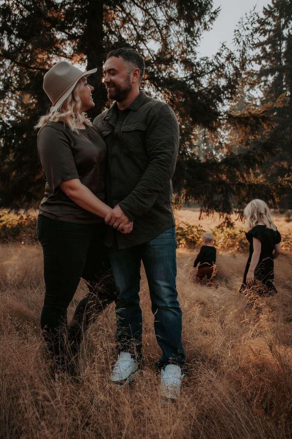A couple in a hat and dark casual clothing holds hands in a tall grass field while two children play in the background at golden hour.