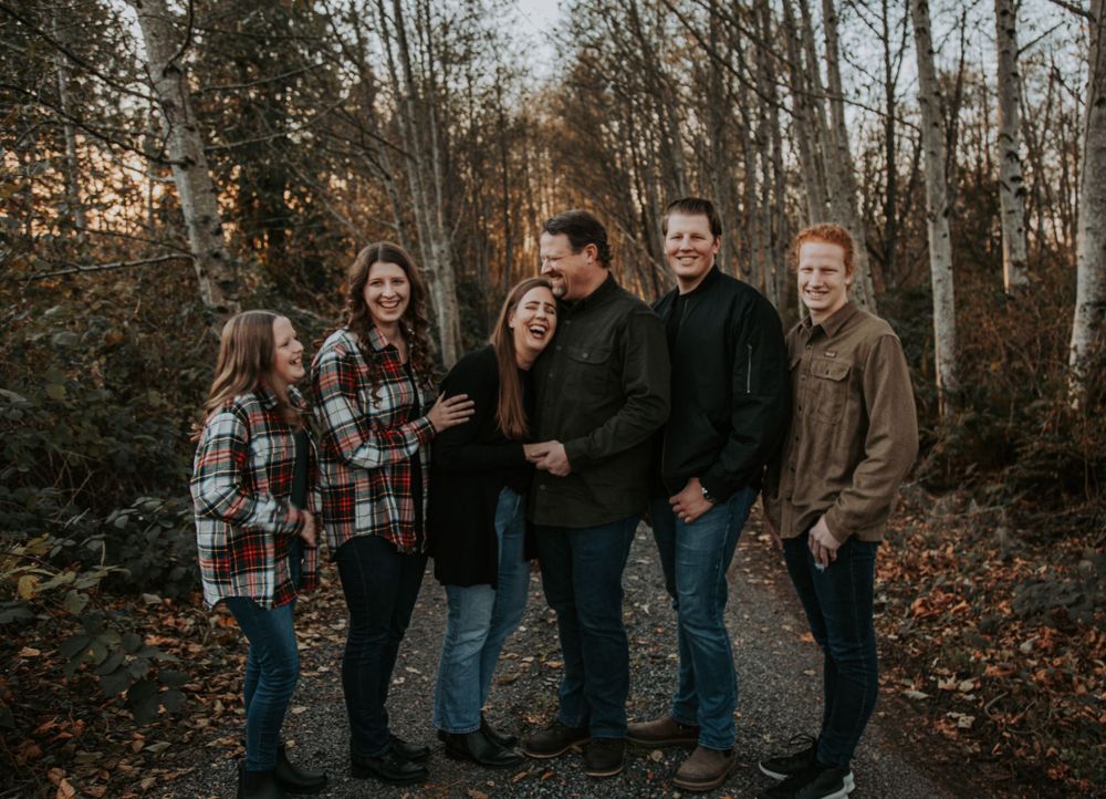 A family of six in flannel and earthy tones stands together on a gravel path lined with birch trees in the fall.