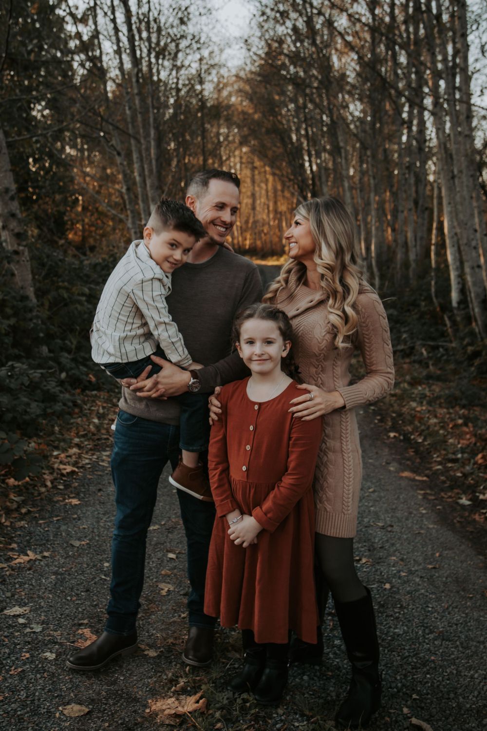 A family of four in warm autumn outfits stands on a leaf-covered forest path, the father holding his son beside the mother and daughter.