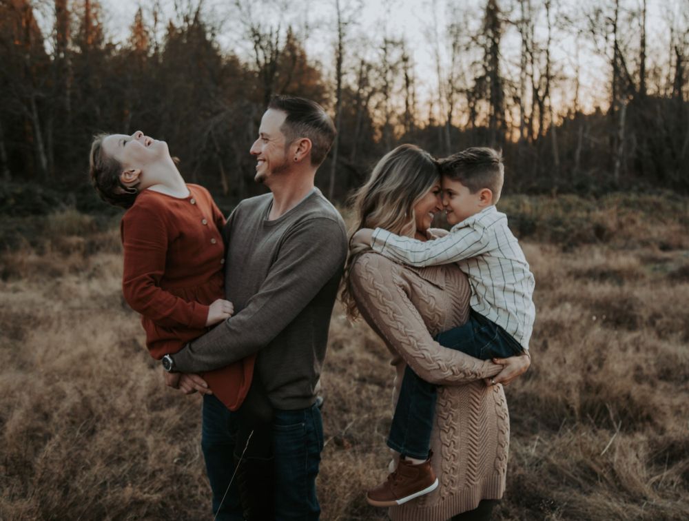 A joyful family laughs and embraces in a golden field at sunset, with parents lifting their two children during a lifestyle photo shoot.