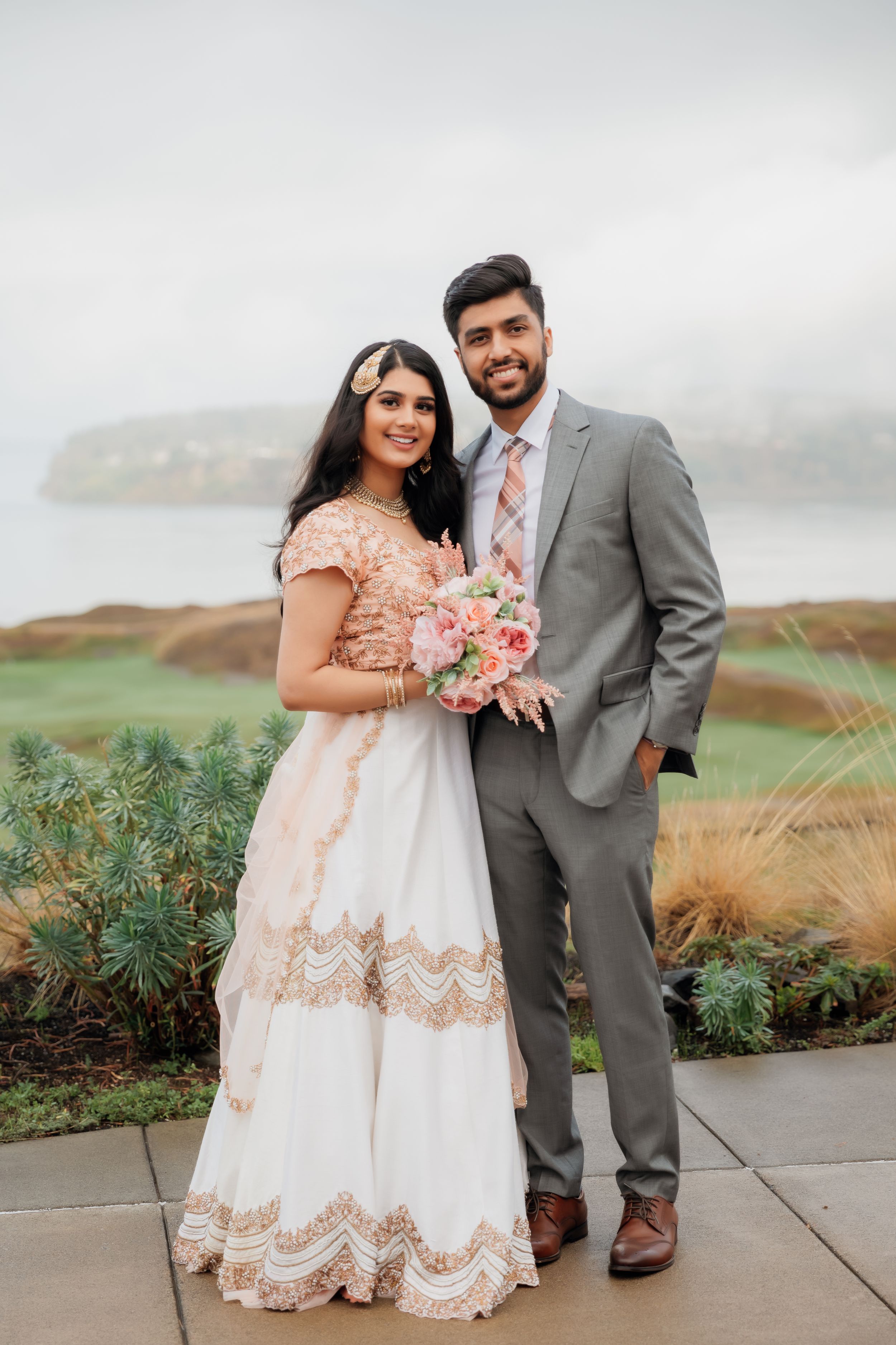 A couple in wedding attire poses outdoors with a scenic coastal backdrop, the bride holding a pink bouquet.