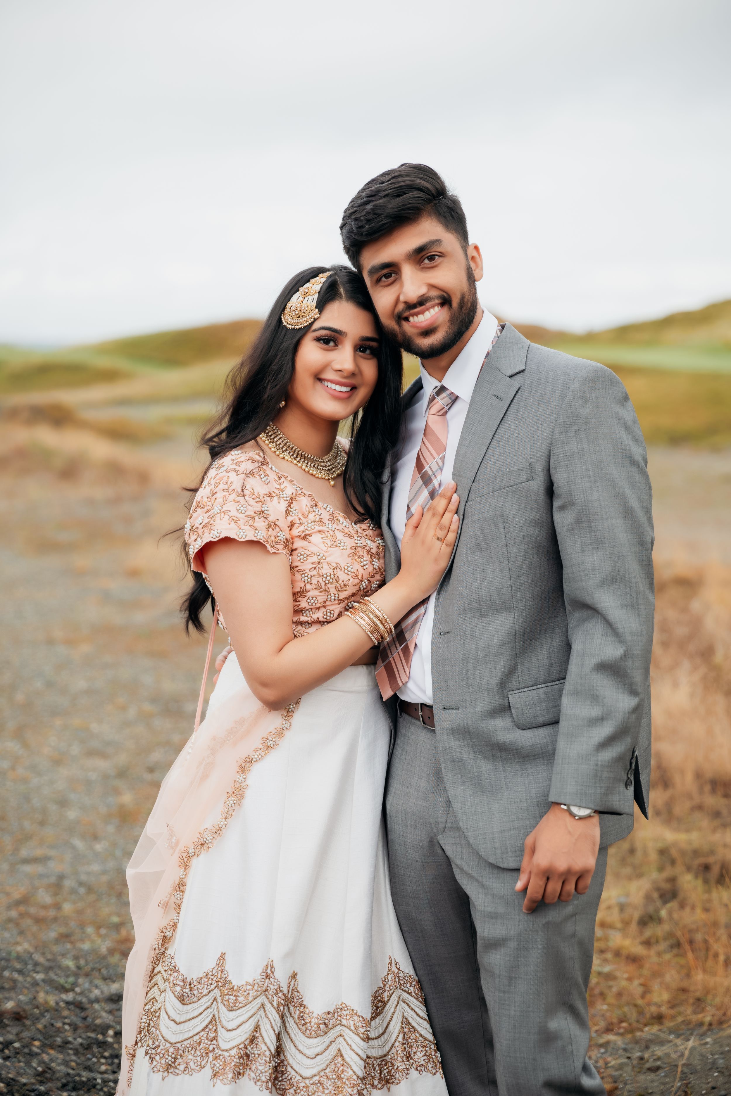 A couple smiles together outdoors, with the bride wearing an ornate white and gold dress.