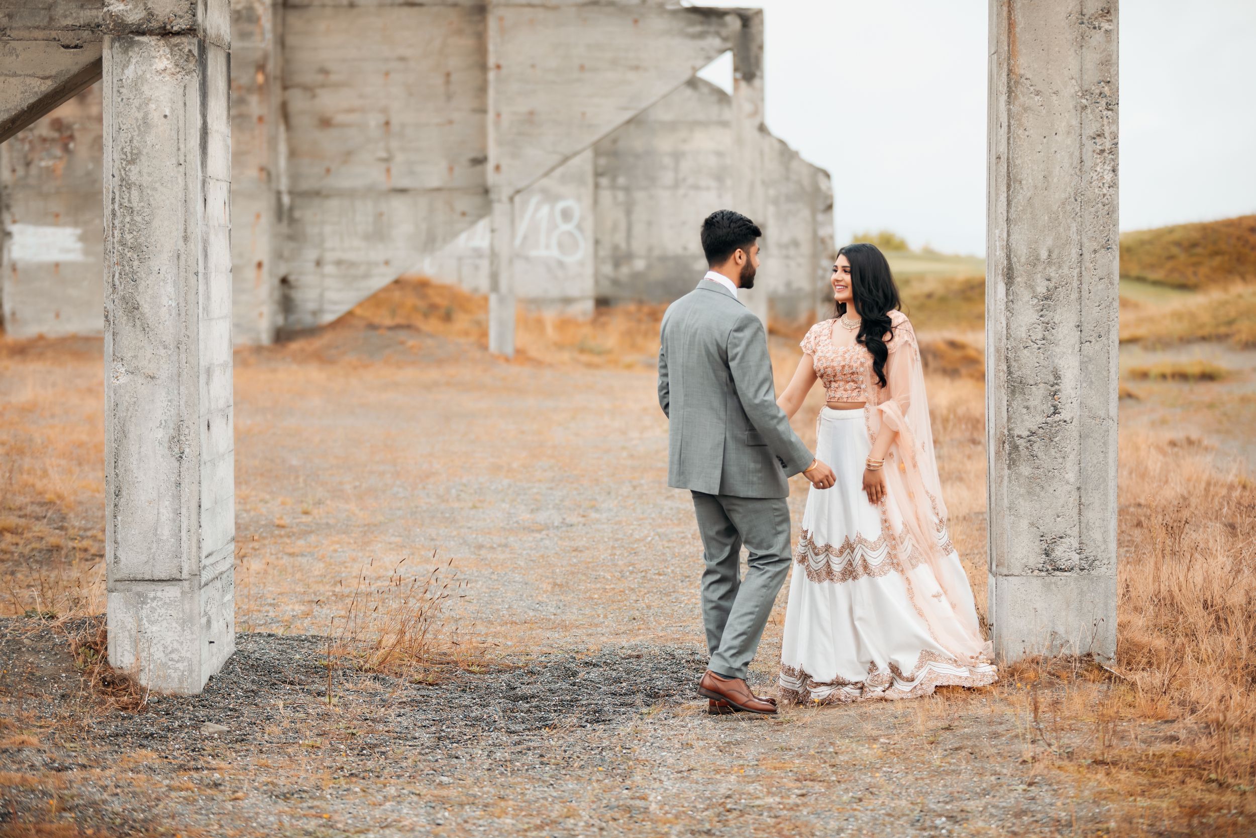 A couple stands between concrete pillars in a rustic outdoor setting wearing formal attire.