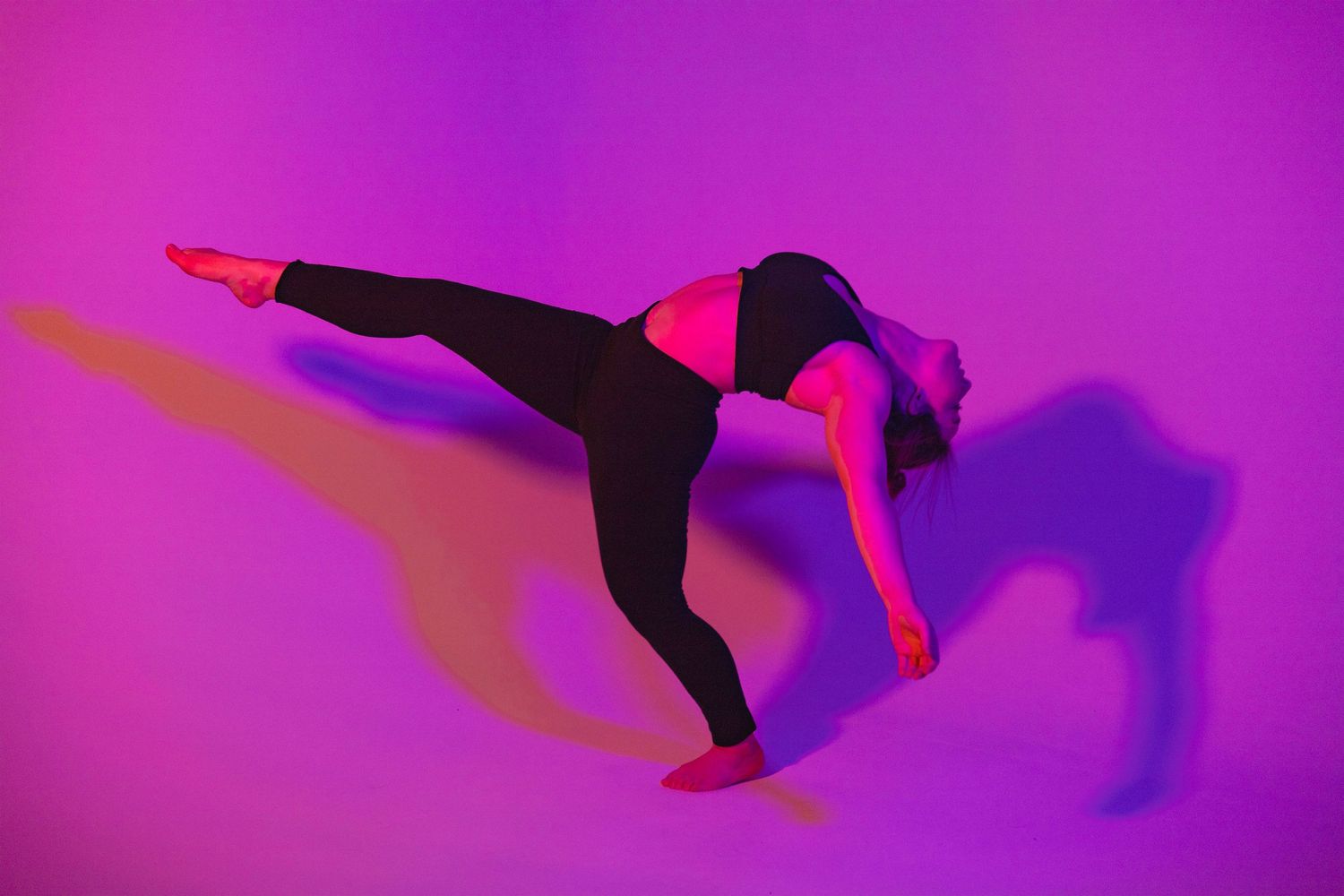 Dancer in black stretches gracefully on floor bathed in vibrant pink and purple lighting.