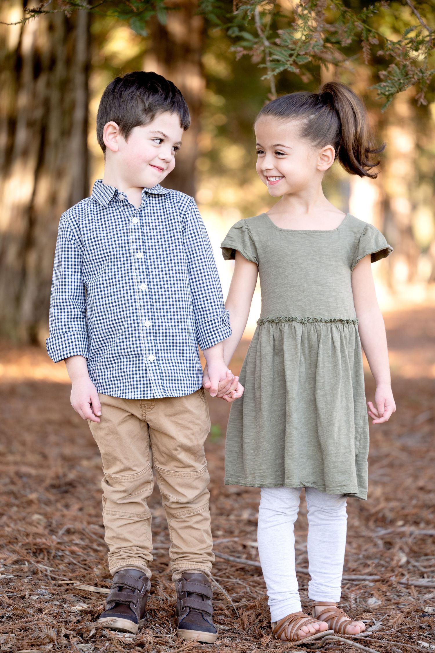 Two children in coordinating outfits hold hands while standing on fallen leaves in a wooded setting.