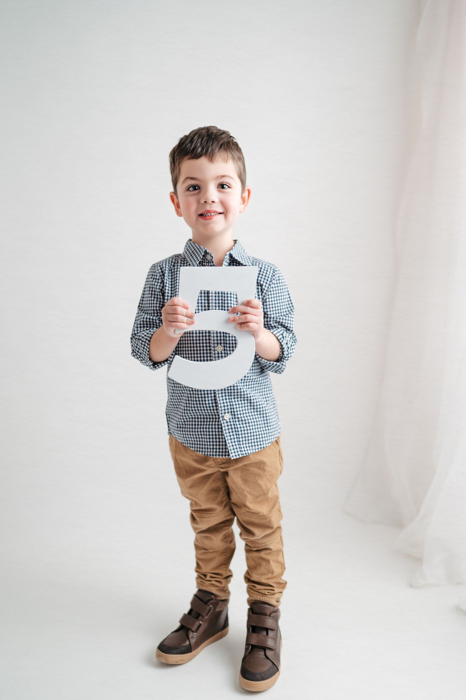 A young boy in a checkered shirt and khakis holds up a decorative number while posing against a white background.