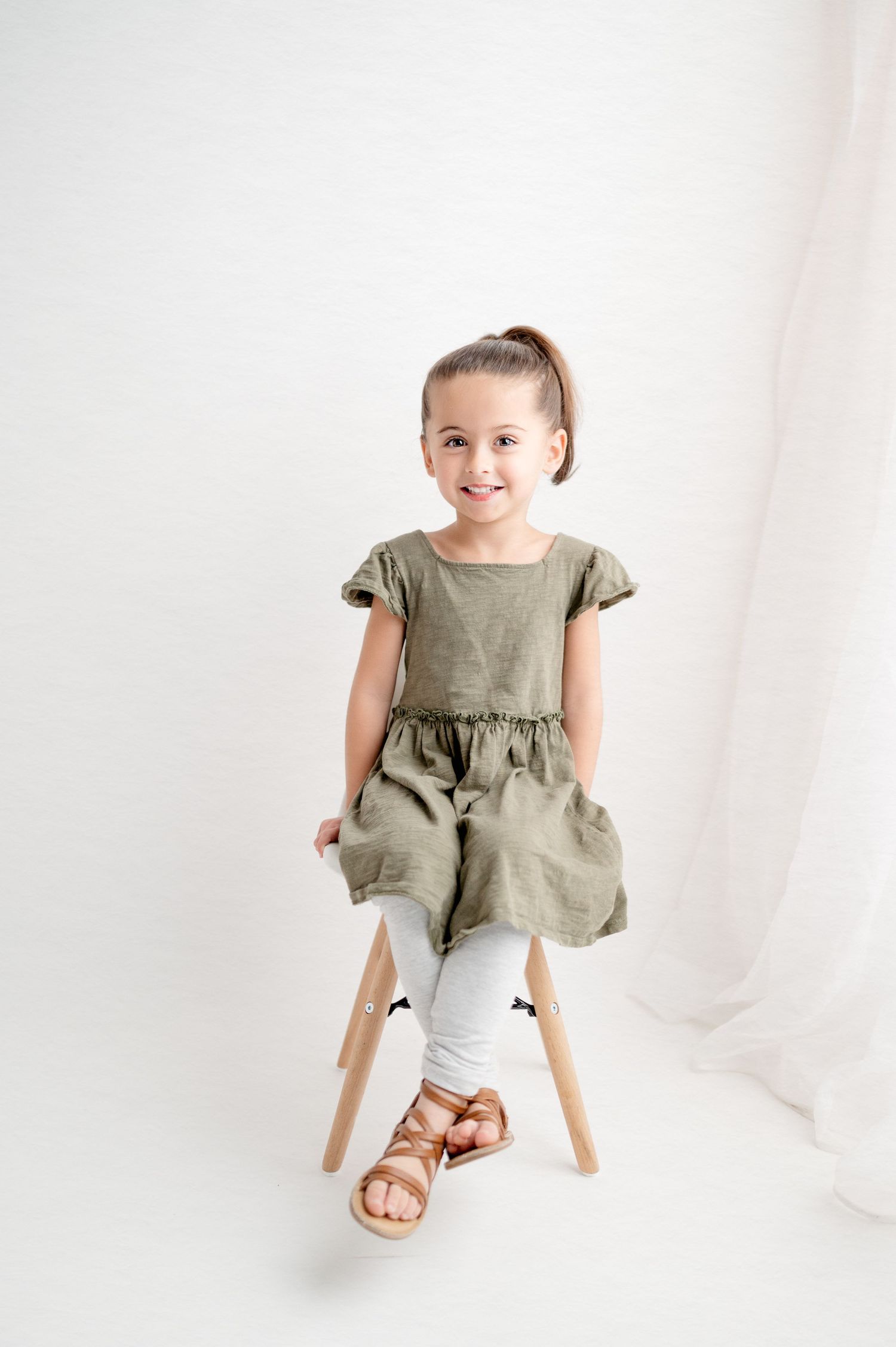 Girl in olive dress sitting on wooden chair against white background.