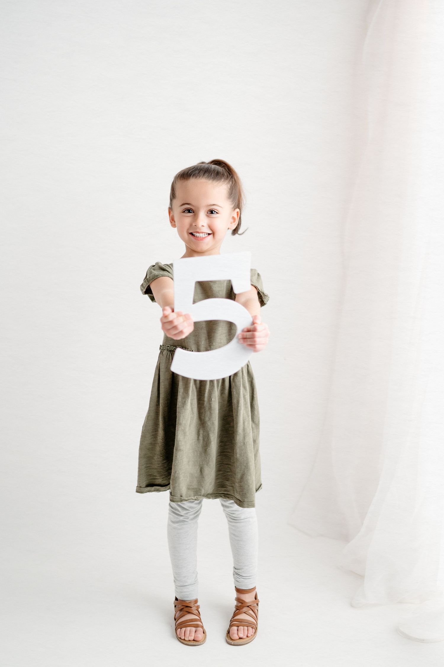 A young girl in an olive dress and white leggings holds up a large decorative letter while standing against a white backdrop.