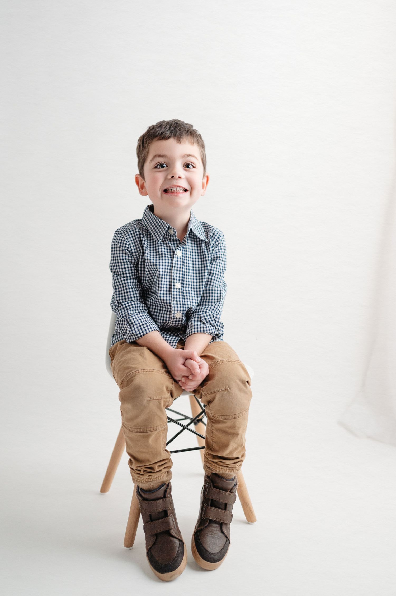 Young boy in checkered shirt and khakis sitting on wooden chair against white background.
