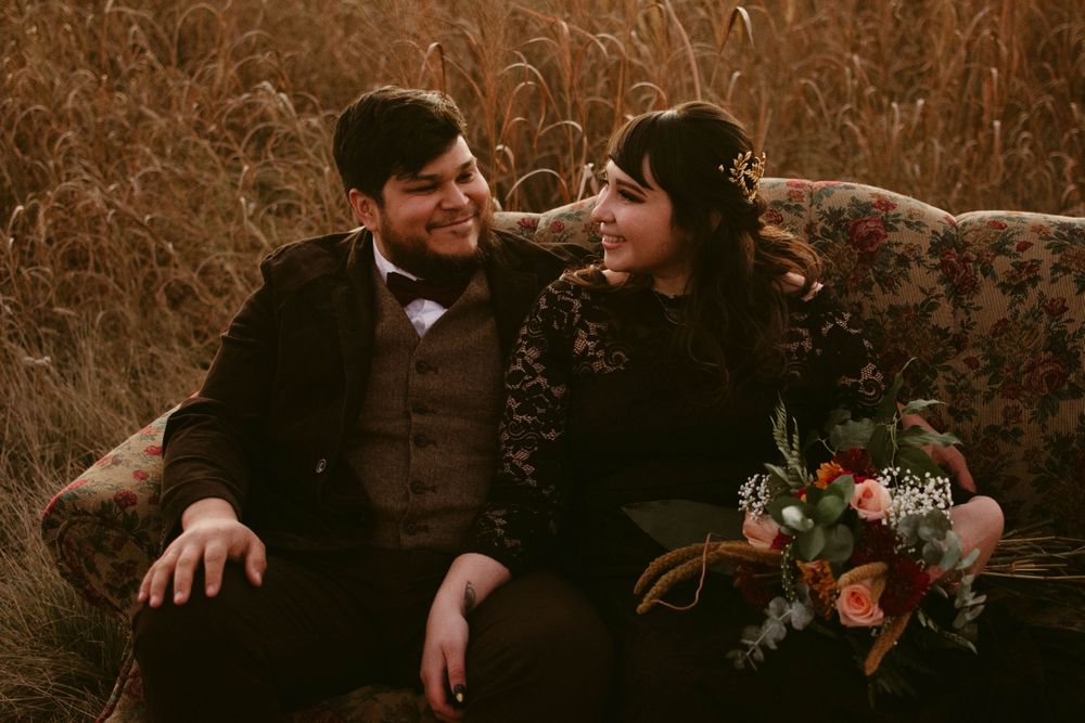 A couple in formal attire shares a tender moment on a vintage couch in a field.