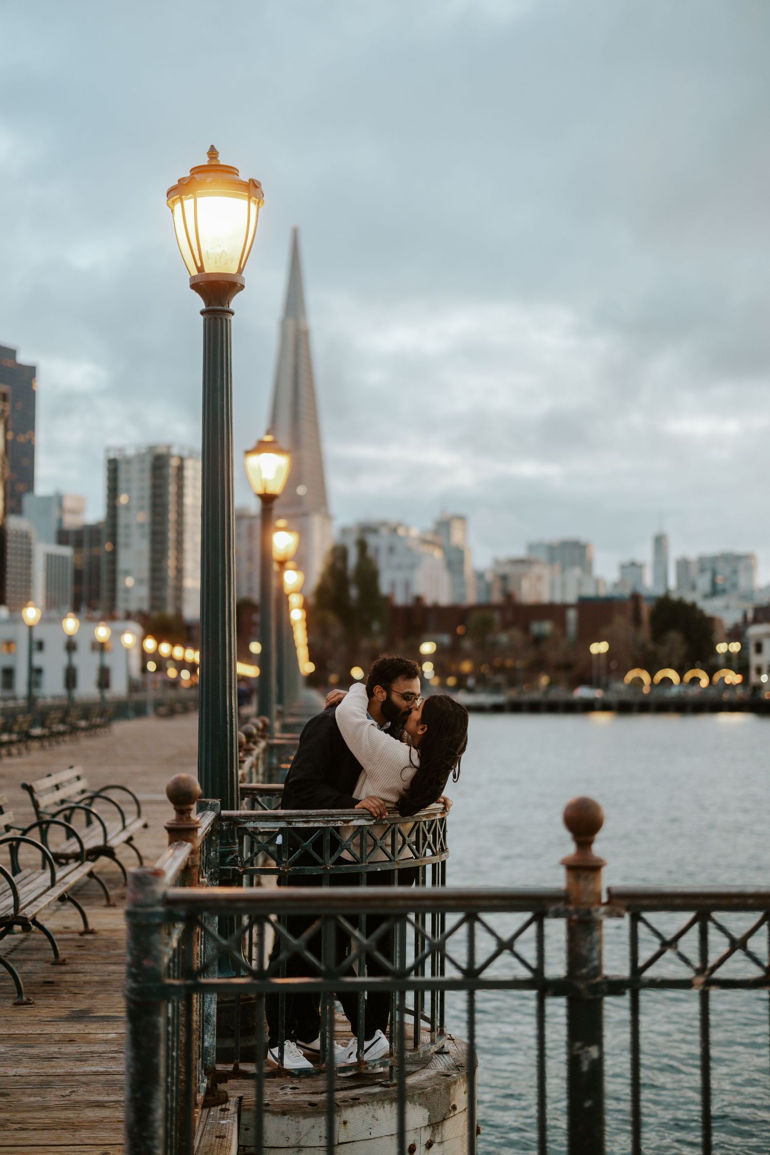 couple kissing and leaning over a railing at Pier 7 with the street lamps on and the the San Francisco skyline behind them during engagement