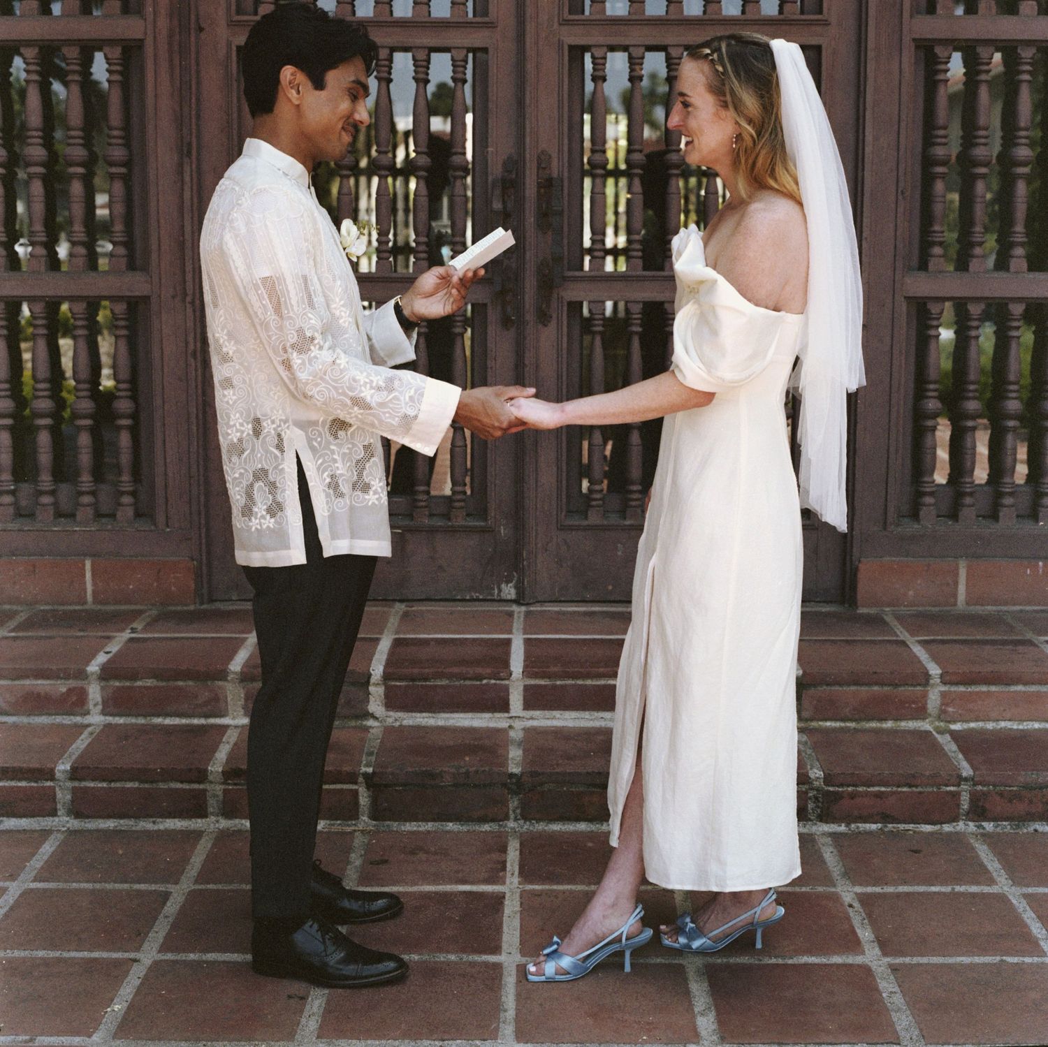 Wedding couple exchanges vows on terracotta tiles in front of ornate wooden gates.