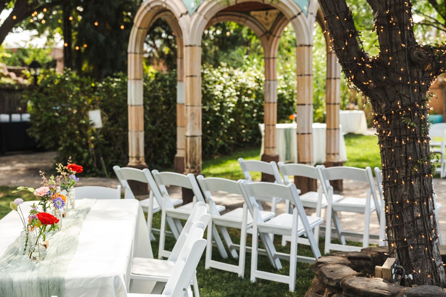 White folding chairs arranged for an outdoor wedding ceremony under an elegant stone archway and tree.
