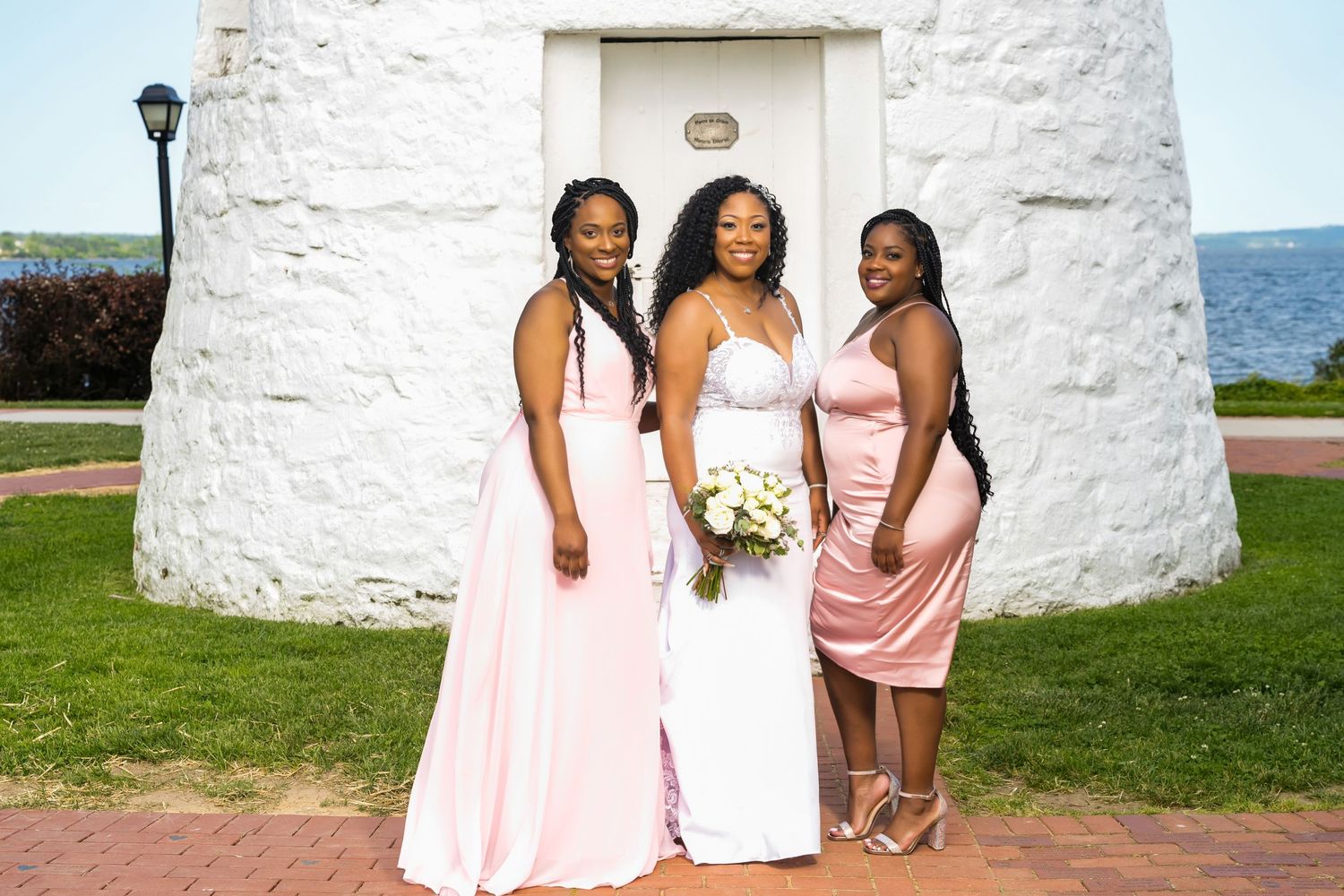 Three women in pink dresses pose in front of a white lighthouse.