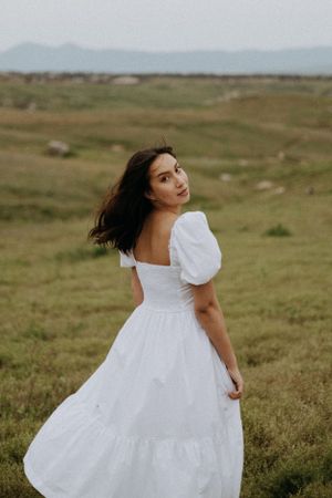 A person in a white puff-sleeve dress poses against rolling hills and mountains.