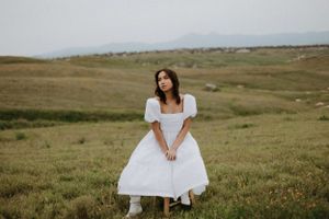 A dreamy portrait of someone in a white dress sitting in a vast meadow landscape.