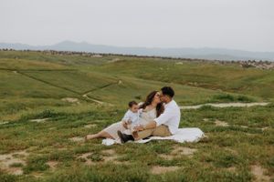 A couple enjoys a picnic on a blanket in a vast green meadow with mountains in the distance.