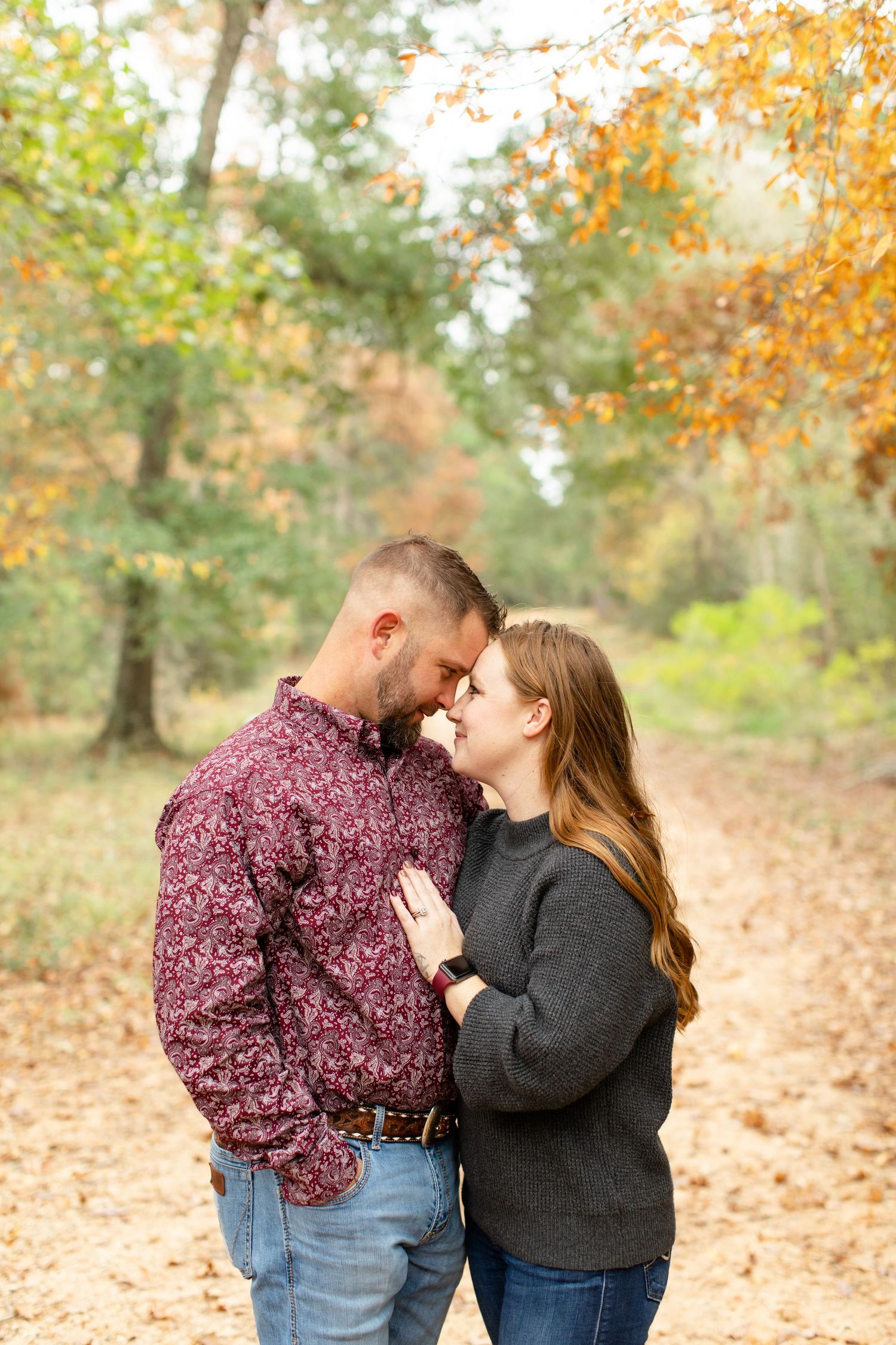 Couple embracing on leaf-covered trail with golden autumn trees in background.