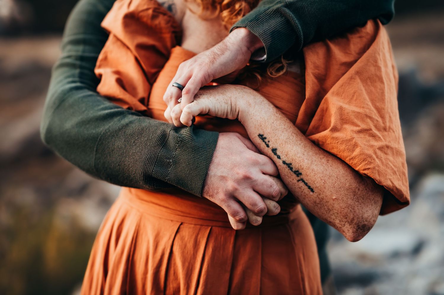 close up of arms of husband in green sweater wrapping around wife in orange dress for a hug.