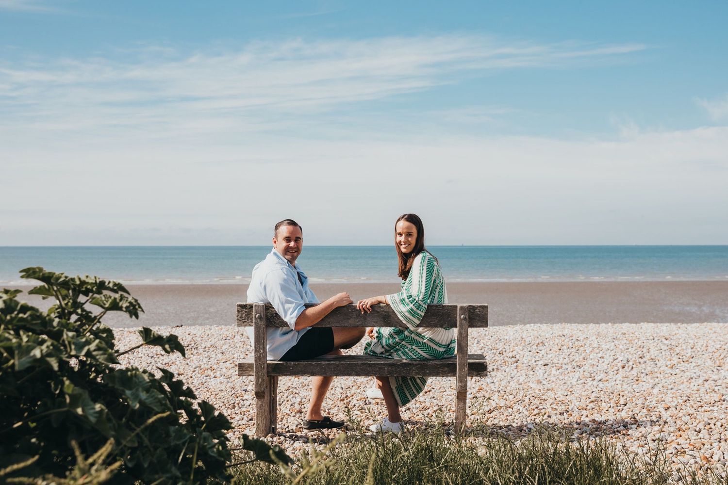 A couple sits together on a seaside bench with a pebble beach and ocean view behind them.