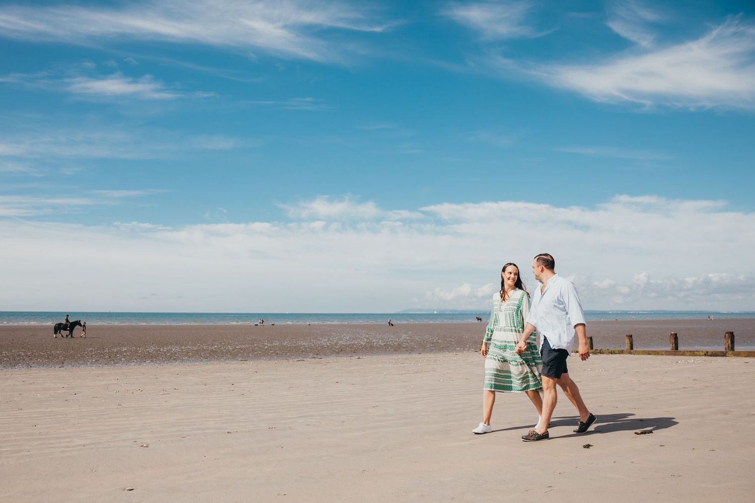 Two people stroll along a vast empty beach with blue skies and a horse in the distance.