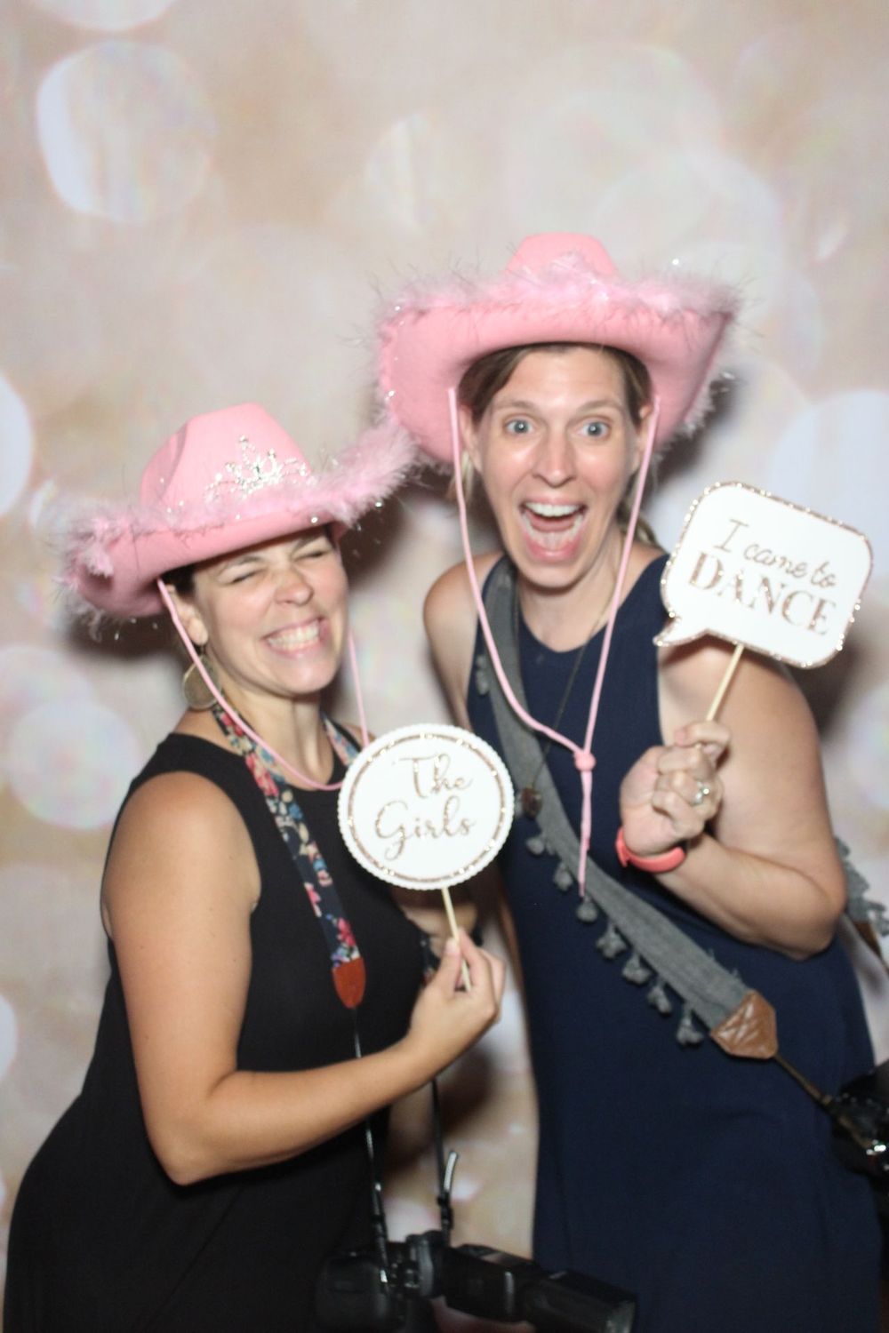 Two people in black outfits pose with pink cowboy hats and photo props against a sparkly backdrop.