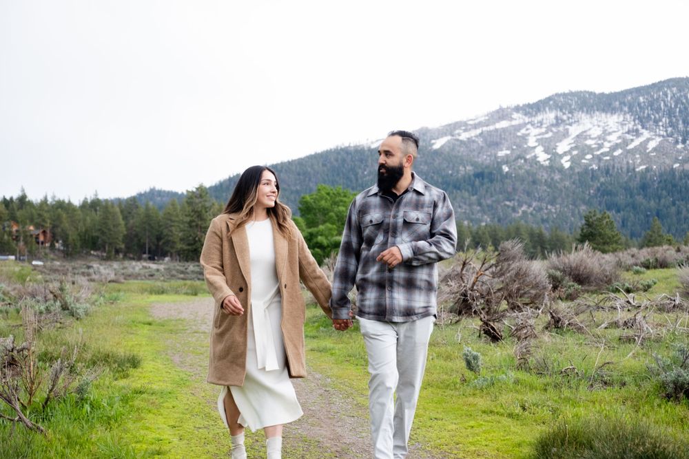 A happy couple take a scenic walk along a mountain trail on a spring day with snow-capped peaks in the background.