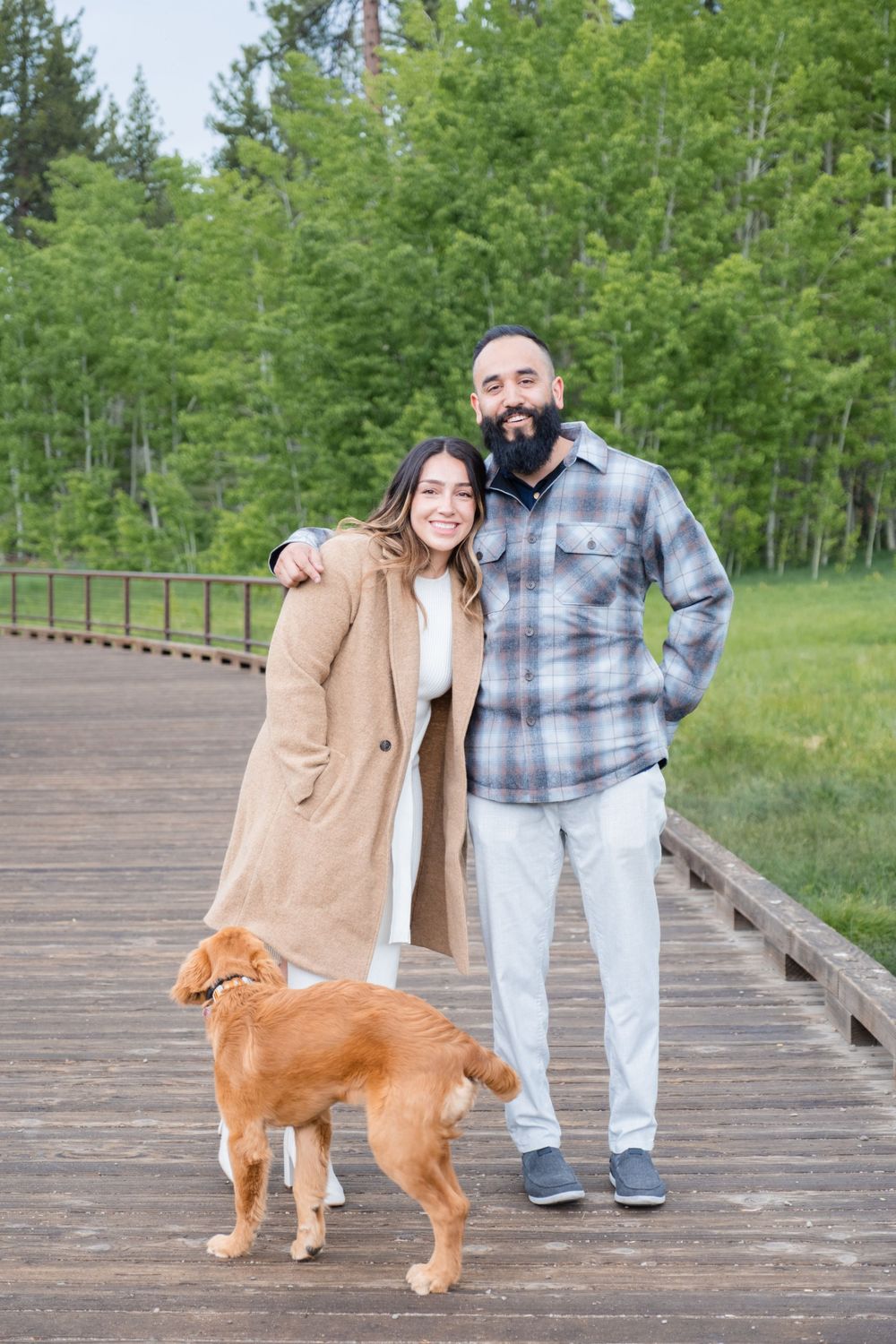 Couple and their golden retriever dog standing together on wooden boardwalk with green forest in background.