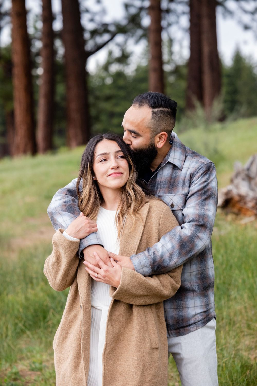 A loving couple embraces outdoors in a forest setting with tall pine trees in the background.