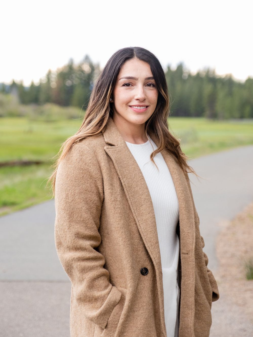 young woman in a cozy camel coat and white sweater stands smiling on a path with evergreen trees in the background.