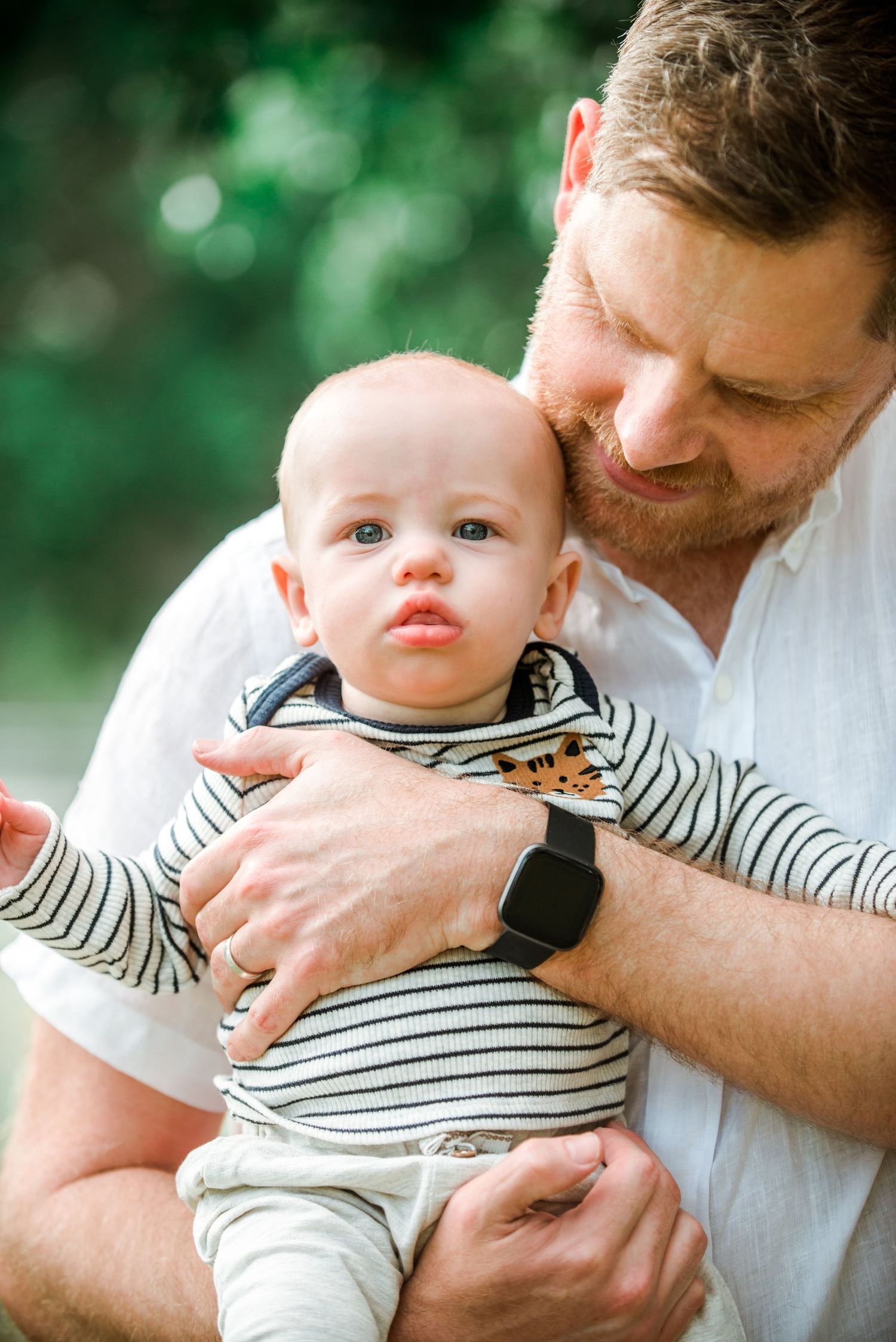 Parent wearing a white shirt holds a baby in a striped outfit against a blurred green background.
