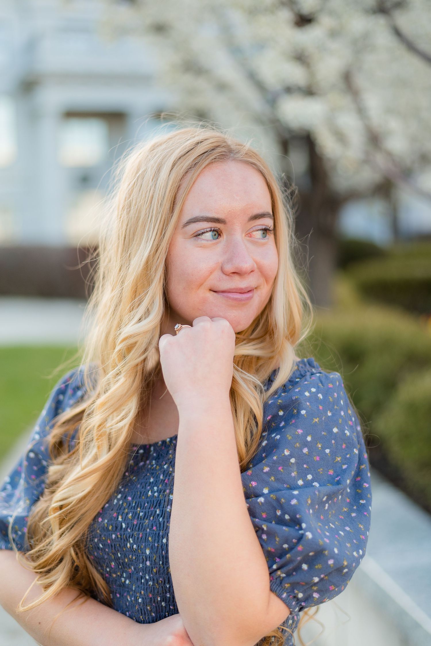 Blonde teenage girl in blue floral dress posing outdoors near blooming trees for senior photos.