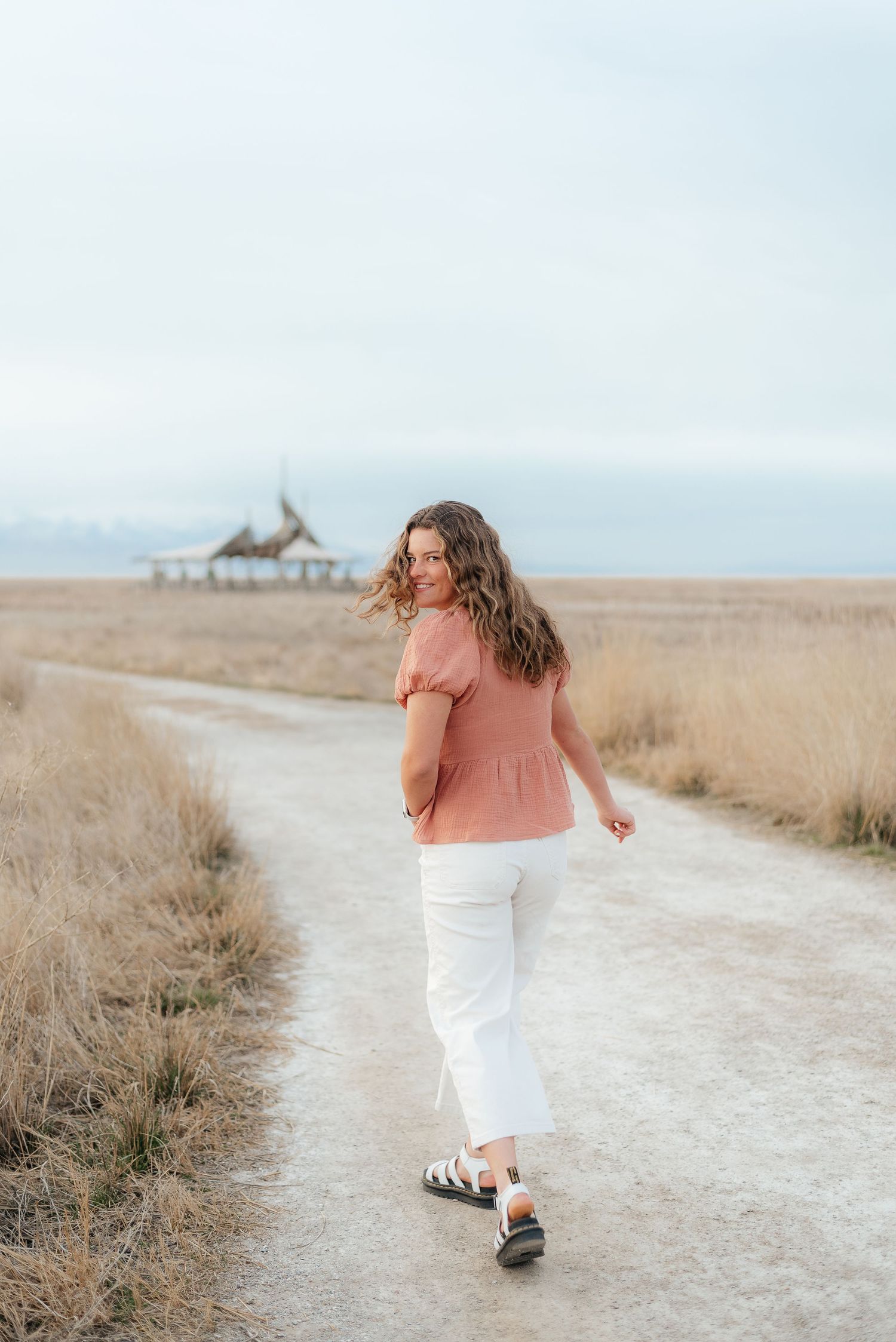 Young woman in pink top and white pants walking on a sandy path through golden fields near the coast.