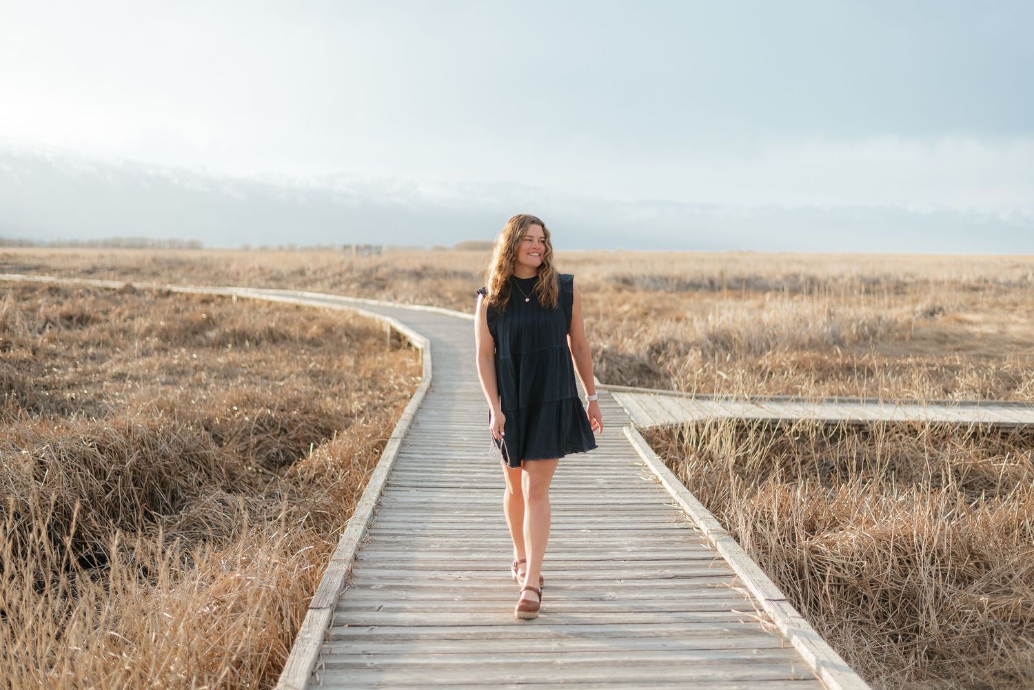 Young woman in black dress walking on a wooden boardwalk through golden fields under a cloudy sky.