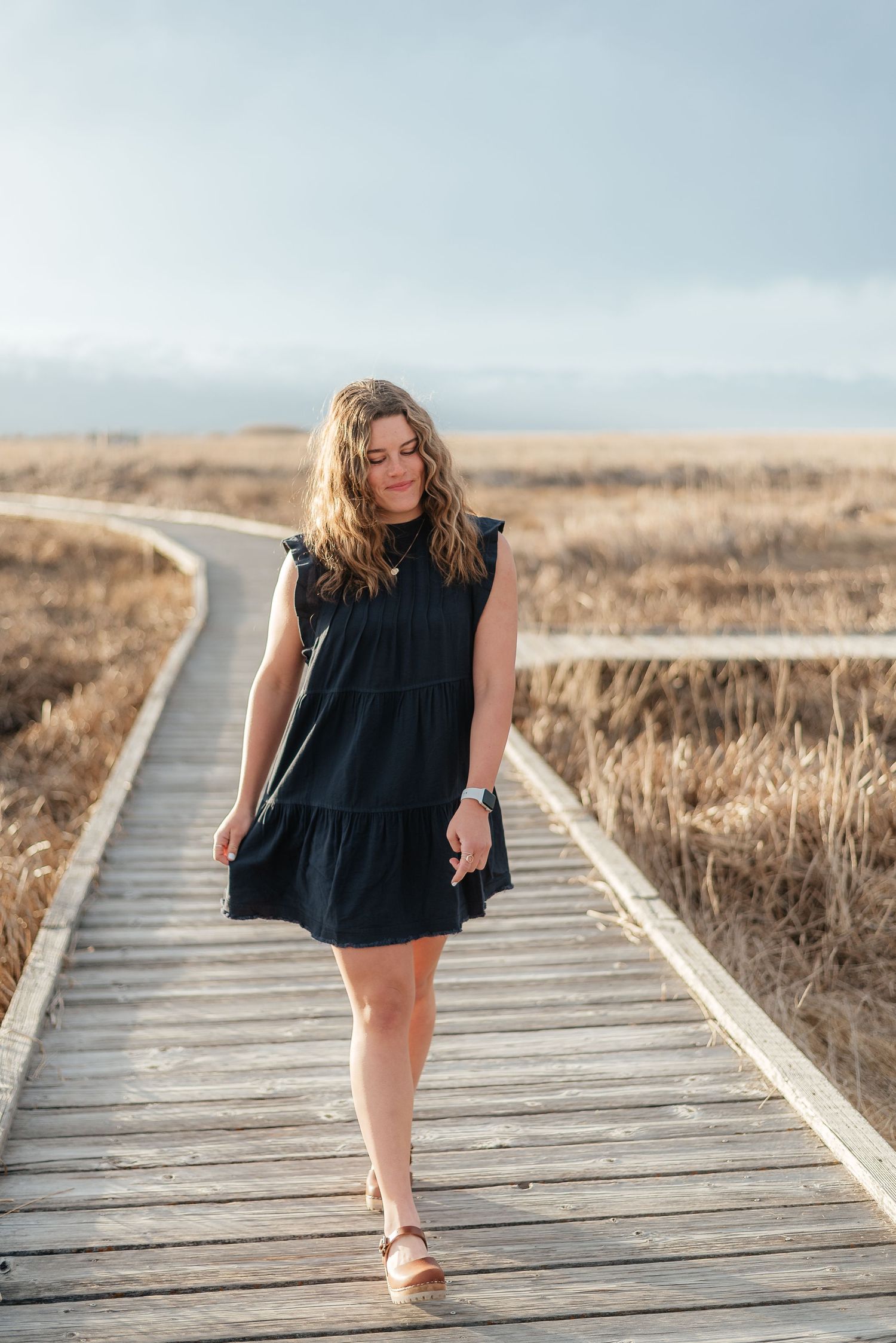 Woman in black sleeveless dress walking on a wooden boardwalk surrounded by dry golden marsh grass.