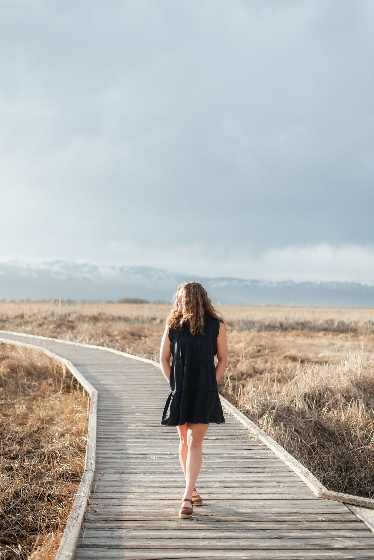 Woman in black dress walking away down a long wooden boardwalk through golden fields toward snowy mountains.