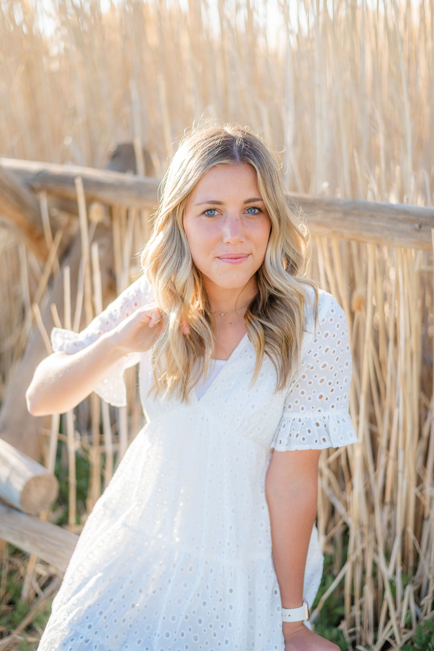 Blonde girl in white eyelet dress standing near rustic wooden fence and golden dry grasses. Megan Snell Photo Utah Weddings