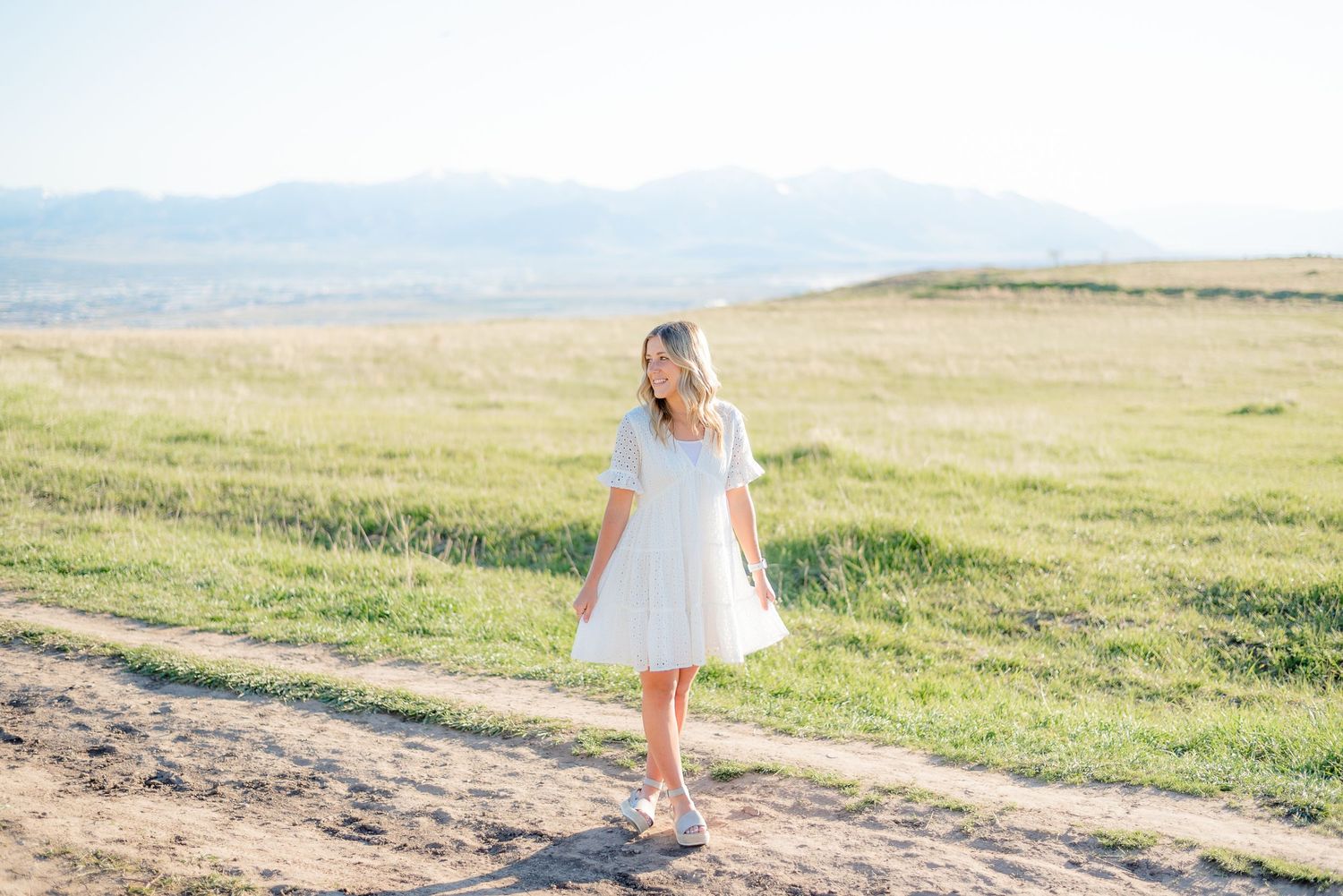 Young blonde woman in white dress walking on dirt path with green fields and mountains in background. Megan Snell Photo Utah Weddings